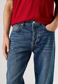 Vaqueros de mezclilla azul con corte recto, que presentan una etiqueta detallada en el bolsillo trasero, cierre con botón metálico y un sutil desvanecimiento a lo largo de la tela.