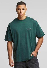 Zaļš kokvilnas T-krekls ar apaļu kaklu un īsām piedurknēm, ar baltu logo druku kreisajā krūtīs. Ikdienas piegriezums.