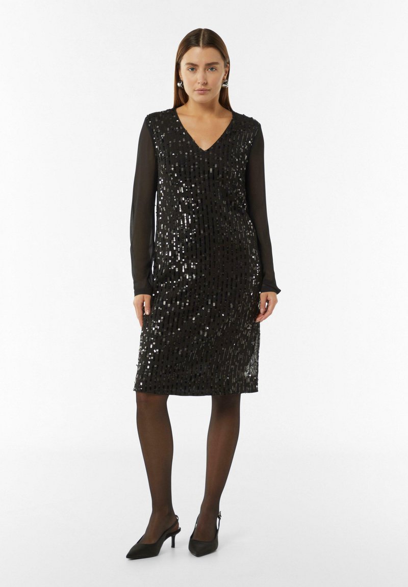 Czarna sukienka z cekinami, z dekoltem w kształcie litery V i długimi, przezroczystymi rękawami, długość do kolan, o dopasowanej sylwetce, zestawiona z czarnymi szpilkami.