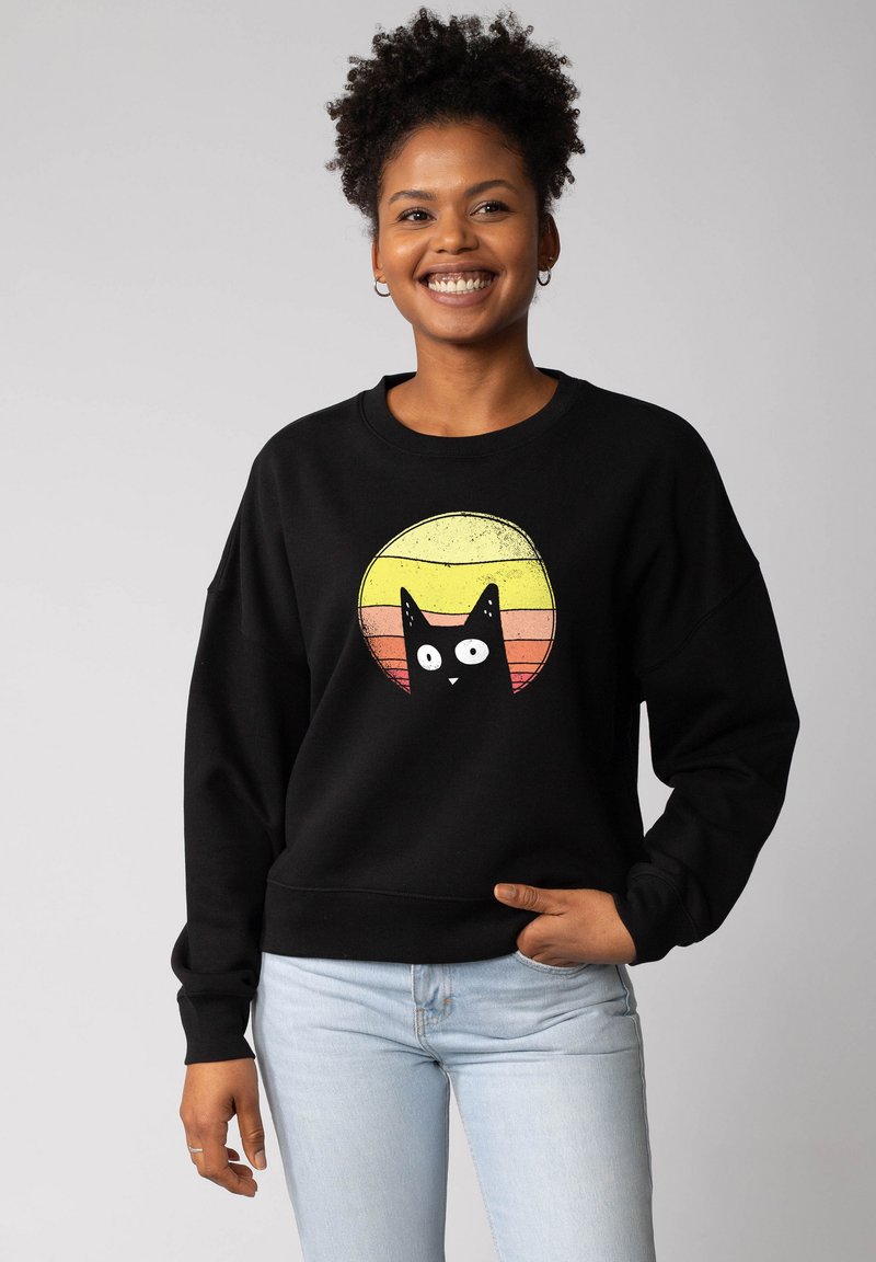 Sweatshirt noir à coupe oversize ; présente un graphisme d'un chat en dessin animé sur un fond circulaire jaune et rose. Texture douce.