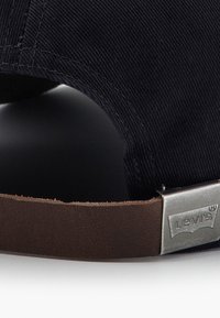 Crna kačket od manšestera s smeđom kožnom trakom s metalnom srebrnom kopčom ugraviranom s "Levi's". Prikaz izbliza materijala i teksture.