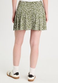 Jupe plissée verte avec un motif floral blanc. Fabriquée en tissu léger, elle dispose d'une taille élastique. Portée avec des chaussures de sport et des chaussettes blanches.