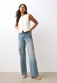 Weißes ärmelloses Hemd mit großen runden Knöpfen, kombiniert mit ausgewaschenen blauen Jeans mit weitem Bein und hellrosa Schuhen mit klobigem Absatz.