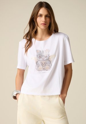 Kvinde iført hvid t-shirt med et palietbesat kattegraphic og cremefarvede bukser, hænderne i lommerne, mod en beige baggrund.