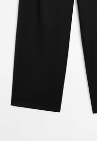 Pantalon noir à jambe droite posé à plat sur une surface blanche, montrant la partie inférieure des deux jambes.