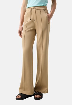 Osoba nosząca beżowe spodnie o szerokich nogawkach z przednimi szwami, elastycznym pasem z sznurkiem, kieszeniami po bokach oraz czarne sandały z plecionymi paskami.