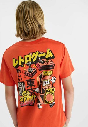 Personne aux cheveux blonds portant un t-shirt orange arborant des graphismes colorés de jeux vidéo rétro et du texte japonais au dos.