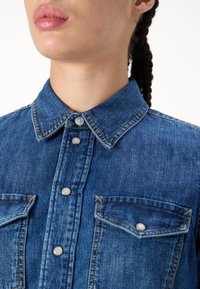 Camicia di denim con colletto button-down, due tasche sul petto e dettagli in metallo argentato. Presenta un colore blu sbiadito e cuciture visibili.