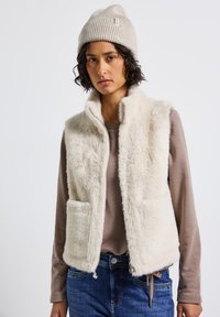 Gilet in finto pelliccia color crema con colletto rialzato, due tasche frontali e superficie texture; abbinato a una camicia a maniche lunghe leggermente beige.
