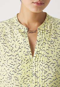 Gul bluse med svarte abstrakte mønstre, plisserte detaljer ved halsen og et smykke med teksten "Karl." Myk tekstur.