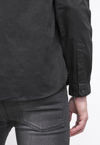 Camicia nera con bottoni, dal tessuto liscio e maniche lunghe, mostrata di schiena, abbinata a jeans grigio scuro.