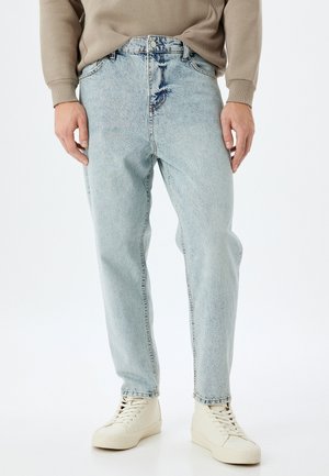 Vaqueros de mezclilla azul claro con un corte holgado, que presentan un lavado desvanecido y un ligero desgaste cerca de la cintura, combinados con zapatillas blancas.