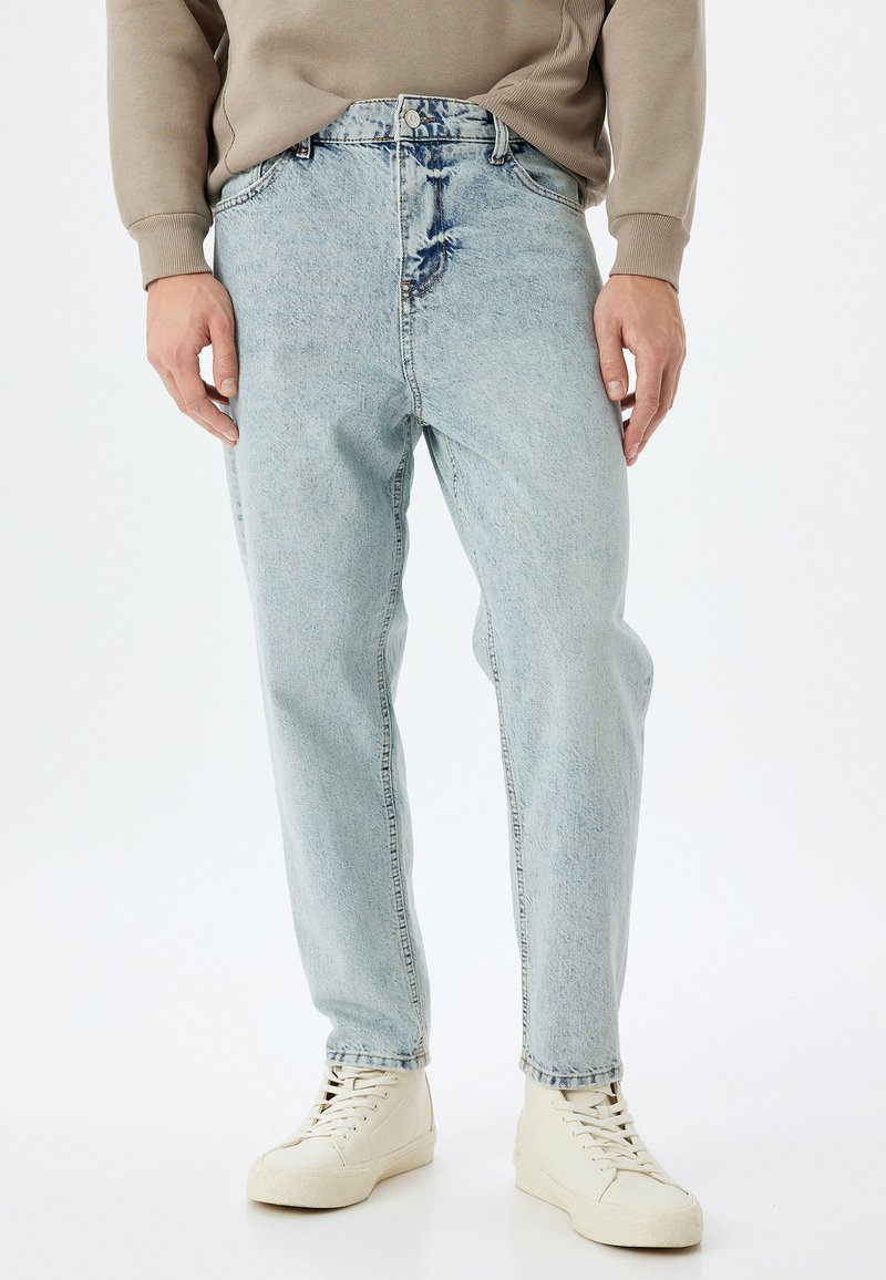 Jeans in denim azzurro chiaro con una vestibilità comoda, caratterizzati da un lavaggio sbiadito e da leggeri segni di usura vicino all'elastico in vita, abbinati a sneakers bianche.