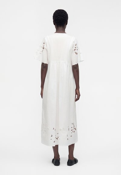 Personne debout de dos portant une robe midi blanche avec des découpes florales sur les manches et l'ourlet, associée à des chaussures plates noires.