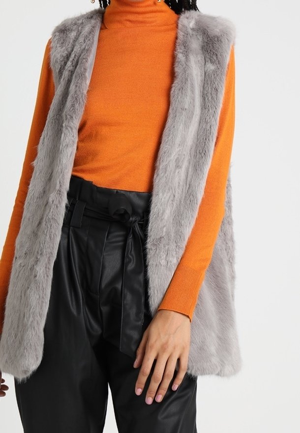 Gilet in ecopelliccia grigia indossato su un dolcevita arancione, abbinato a pantaloni neri a vita alta con cintura annodata, che mostrano texture diverse.