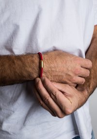Raudona pintinė apyrankė su auksiniu cilindriniu užsegimu, dėvima ant rankos, turinti struktūruotą paviršių ir kontrastingas spalvas.