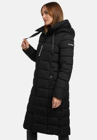 Manteau matelassé noir long avec capuche, doté d'une fermeture éclair à l'avant, de zips sur les côtés, d'un design matelassé et de quincaillerie de marque sur la manche.