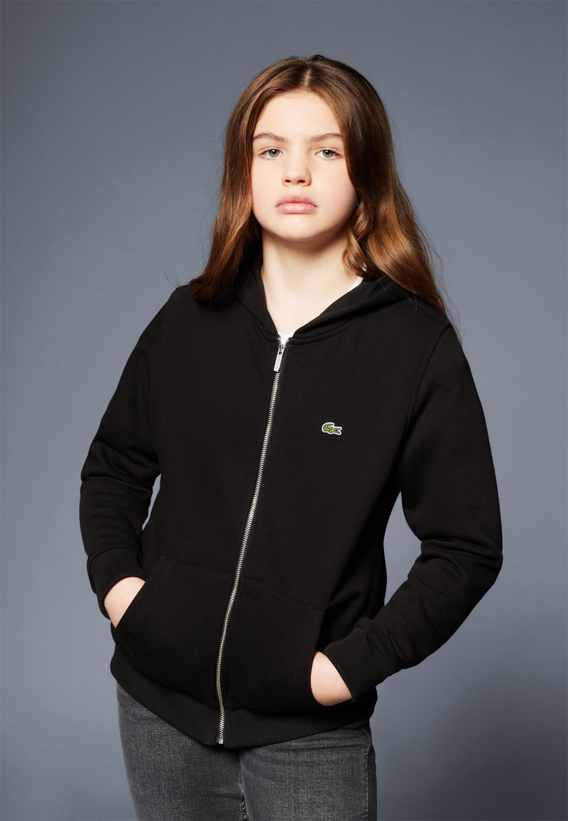 Černá mikina na zip s klokaní kapsou, žebrovanými manžetami a malým zeleným logem Lacoste na levé hrudi. Nosené s šedými džíny.