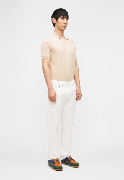 Άνδρας που στέκεται μπροστά, φοράει μπεζ πόλο πουκάμισο, λευκό παντελόνι και πολύχρωμα καφέ, πράσινα και μπλε παπούτσια με κορδόνια σε λευκό φόντο.