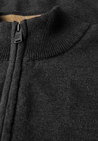 Černý svetr na zip s vroubkovaným výstřihem a texturou. Obsahuje výrazný štítek na zatahovací šňůrce a kontrastní vnitřní límec.