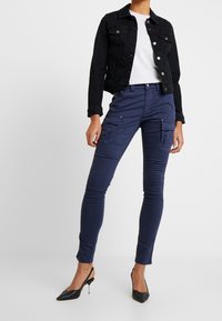 Czarna kurtka dżinsowa z srebrnymi guzikami, noszona na szarej koszulce, w połączeniu z granatowymi spodniami cargo z kieszeniami bocznymi i dopasowanym krojem.