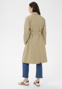 Femme debout, de dos, portant un trench-coat beige, un jean court bleu et des baskets blanches avec des rayures noires.