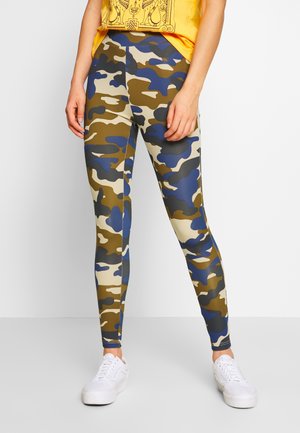 Persoon in groene, beige, blauwe en zwarte camouflageleggings met witte sneakers en een geel patroon top, staande tegen een lichte achtergrond.