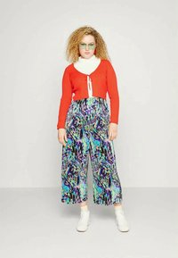 Cardigan cropped riscado vermelho com um suéter de gola alta branco, combinado com calças largas azuis e roxas com padrão. O modelo usa ténis brancos e óculos.