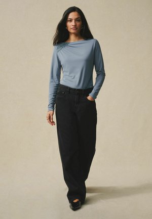 Woman with long dark hair wearing a blue long-sleeve top and wide-leg black pants standing against a plain beige background.