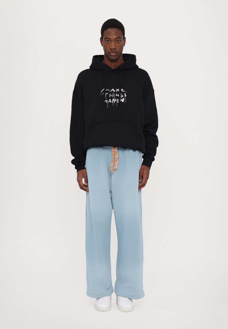 Black hoodie with white graphic text, loose-fit light blue sweatpants, featuring a patterned drawstring, and white sneakers.