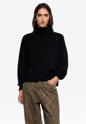 Czarny sweter z golfem o fakturowanej powierzchni i raglanowych rękawach, zestawiony z luźnymi oliwkowozielonymi spodniami z delikatnym wzorem.