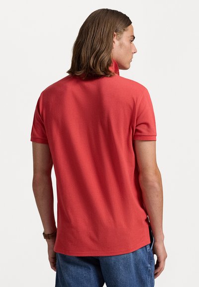 Röd kortärmad polo skjorta i texturerad tyg, med krage och sidslitsar, sedd bakifrån, ihop med blå jeans.
