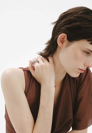 Anillo de plata con acabado texturizado llevado en la mano derecha. El modelo tiene el cabello corto y oscuro y lleva una blusa marrón que revela piel desnuda.