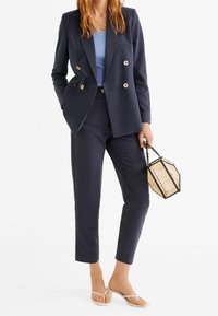 Meriini kaksirivinen bleiseri, yhteensopivat starkatut housut, vaaleansininen paita, läpinäkyvät sandaalit ja geometrinen mustan ja ruskean sävyinen laukku.