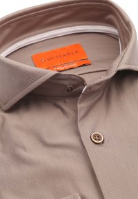 Lichtbruine shirt met een visgraatpatroon, voorzien van een kraag, knoopsluiting aan de voorkant en een oranje label met de tekst "Geschikt voor 4-voudige rek".