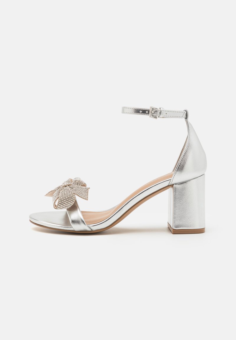 Silver metallic high-heeled sandal with a chunky heel, featuring an embellished bow on the toe strap and ankle buckle closure.