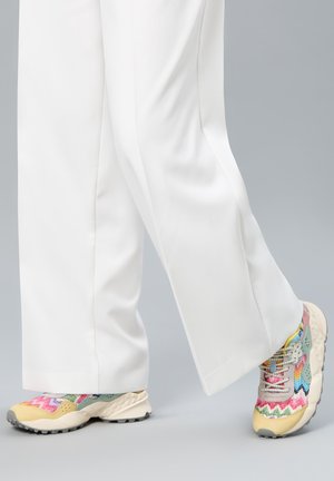 Pantalon blanc à jambes larges avec des baskets à motif zigzag multicolore beige, dotées de semelles épaisses texturées et de lacets, sur un fond gris.