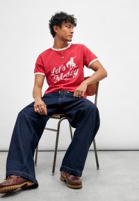 Rød bomull T-skjorte med hvit tekst "La oss mose", parret med mørkeblå vide jeans. Brune sko med lisser, sittende på en metallstol.