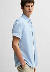 Camicia azzurra a maniche corte in cotone, con colletto a punta, maniche arrotolate e orlo dritto. Leggera texture visibile.