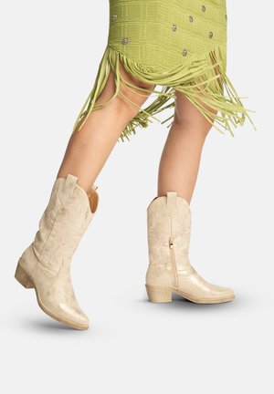 Ben iført lyse beige broderede cowboystøvler kombineret med et grønt frynset skørt, den ene fod løftet midt i et skridt mod en hvid baggrund.