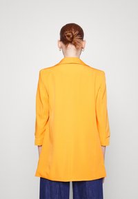 Oranžový blazer s dobře padnoucím střihem, s klopami a tříčtvrtečními rukávy, vyrobený z hladkého materiálu. Pohled zezadu.
