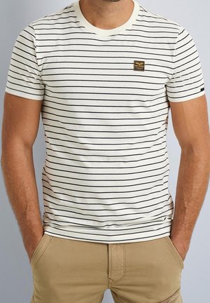 Homme portant un t-shirt blanc à fines rayures noires horizontales et un petit logo sur la poitrine, accompagné d’un pantalon beige, mains dans les poches.