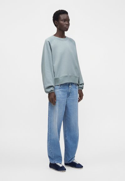 Personne debout portant un sweat à capuche oversize bleu clair, un jean bleu clair ample et des baskets bleu marine avec des lacets blancs.