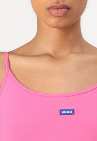 Różowy bezrękawnik z okrągłym dekoltem i cienkimi ramiączkami, z niebieską naszywką "HUGO" przyszytą z przodu. Gładka faktura materiału.