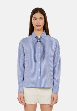 Blauw-witte gestreepte langomshirt met een kraag, met een lintstrik en een sierlijke broche. Draagt een lichte korte broek.