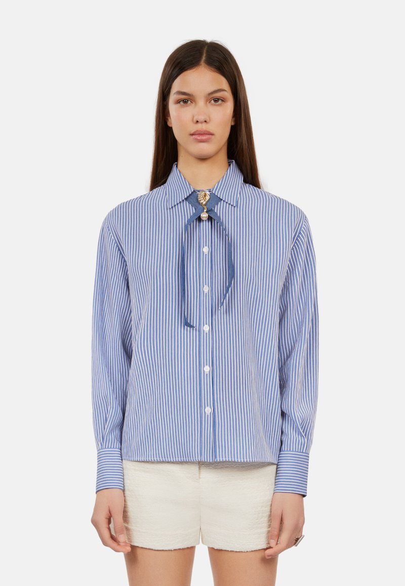 Camicia a maniche lunghe a righe blu e bianche con colletto, caratterizzata da un nastro annodato e una spilla ornamentale. Indossata con pantaloni corti di colore chiaro.