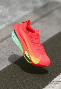 Rød idrettssko med en teksturert overflate, gul swoosh, grønn såle og luftputefunksjon. Lett design, minimale snørebånd.