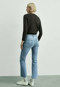Pulover închis la culoare, ajustat, cu gât rotund, asortat cu blugi deschiși la culoare, evazați. Modelul poartă adidași albi și șosete gri, îndreptându-se ușor înapoi.