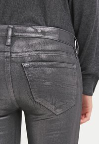 Jeans gris metálico con una textura brillante, que cuentan con un diseño de cinco bolsillos y una pequeña etiqueta de la marca en la cintura.