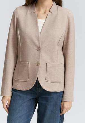 Beiger Zweiknopf-Blazer mit aufgesetzten Fronttaschen, getragen über einem weißen Top, kombiniert mit blauen Jeans an einer stehenden Person.